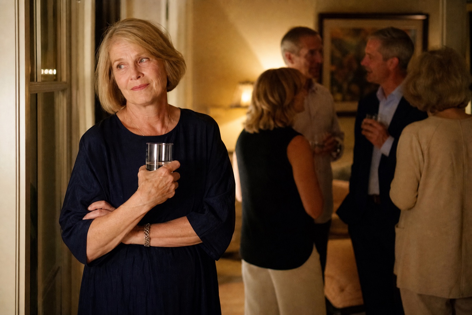 A woman standing slightly apart from the group at a family party, arm crossed over her stomach